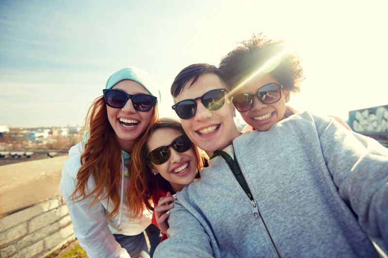 Vier junge Freunde machen draußen bei Sonnenschein fröhlich ein Selfie mit Sonnenbrillen.