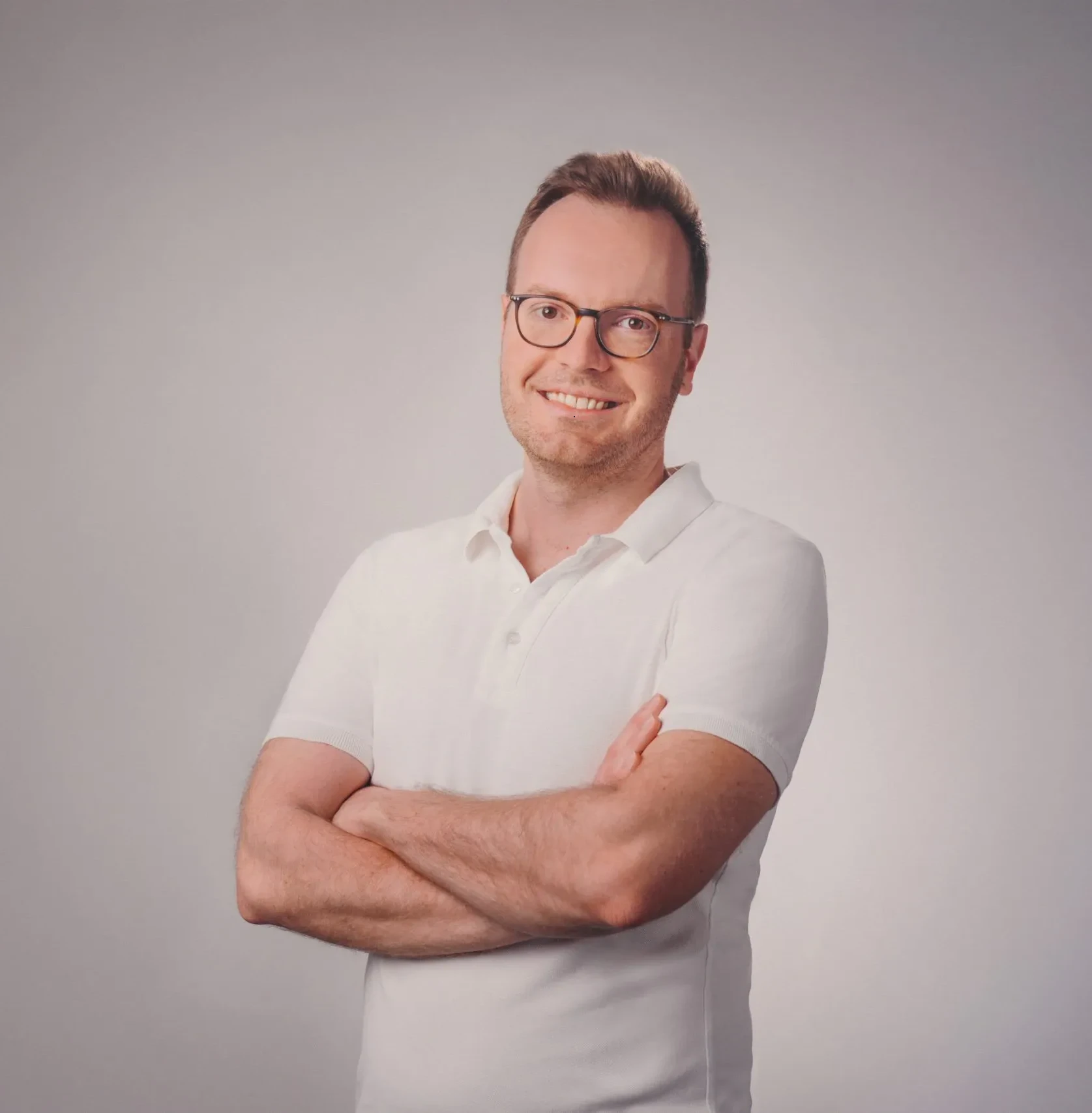 Mann mit Brille und weißem Polohemd lächelt vor grauem Hintergrund, Portraitfoto.